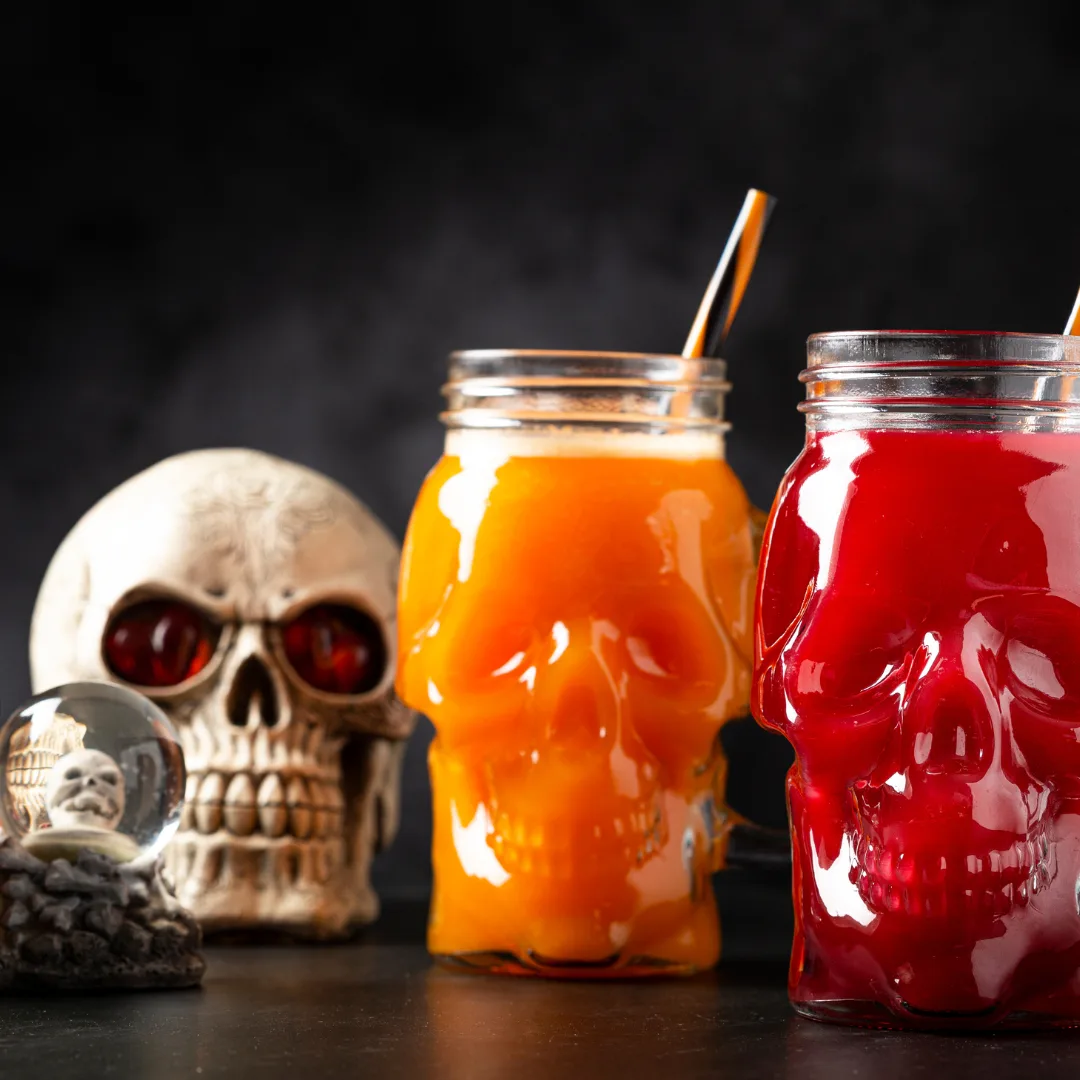 Two Halloween drinks in skull-shaped mugs, one orange and one red, with striped straws, placed beside decorative skulls and a crystal ball. Perfect spooky cocktails for a Halloween party.