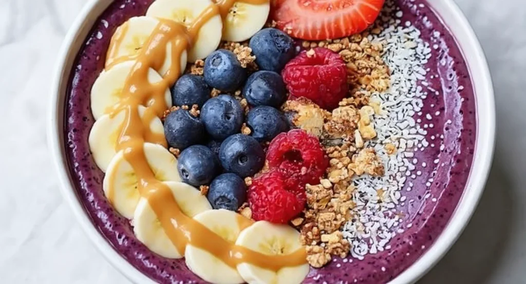 Easy Acai Bowl Recipe served with sliced banana, strawberries, raspberries, blueberries, granola, and a drizzle of peanut butter.