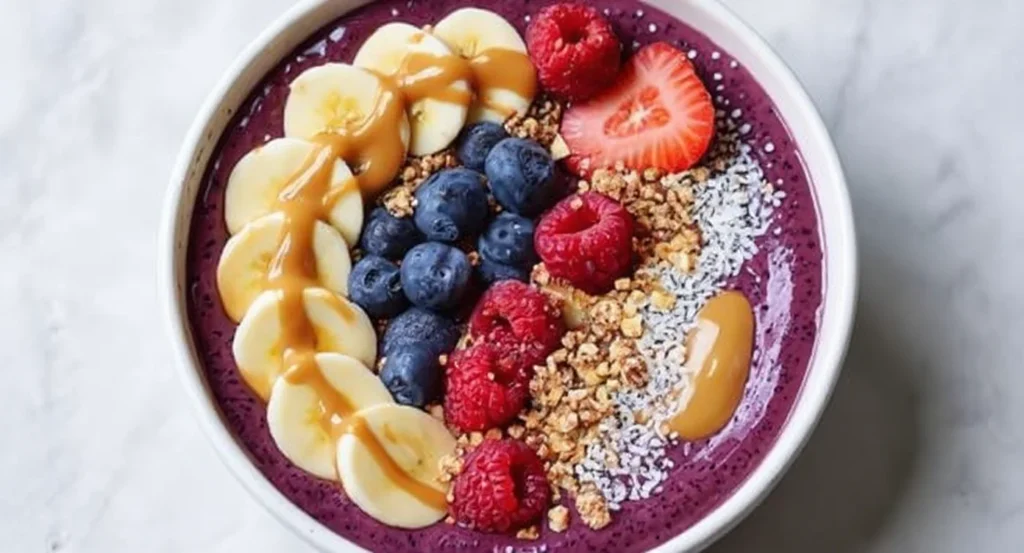 Easy Acai Bowl Recipe topped with banana slices, blueberries, raspberries, strawberries, granola, and peanut butter drizzle.